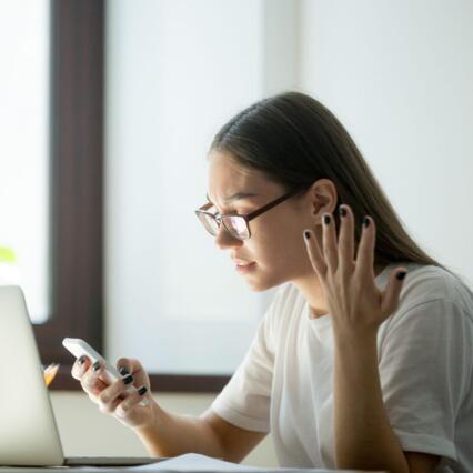 A young woman sitting at a desk looks annoyed at her phone, with one hand thrown up in frustration